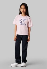 T-shirt in cotone rosa con logo "CK" blu, abbinata a jeans in denim blu scuro e scarpe da ginnastica bianche. Vestibilità comoda e maniche corte.