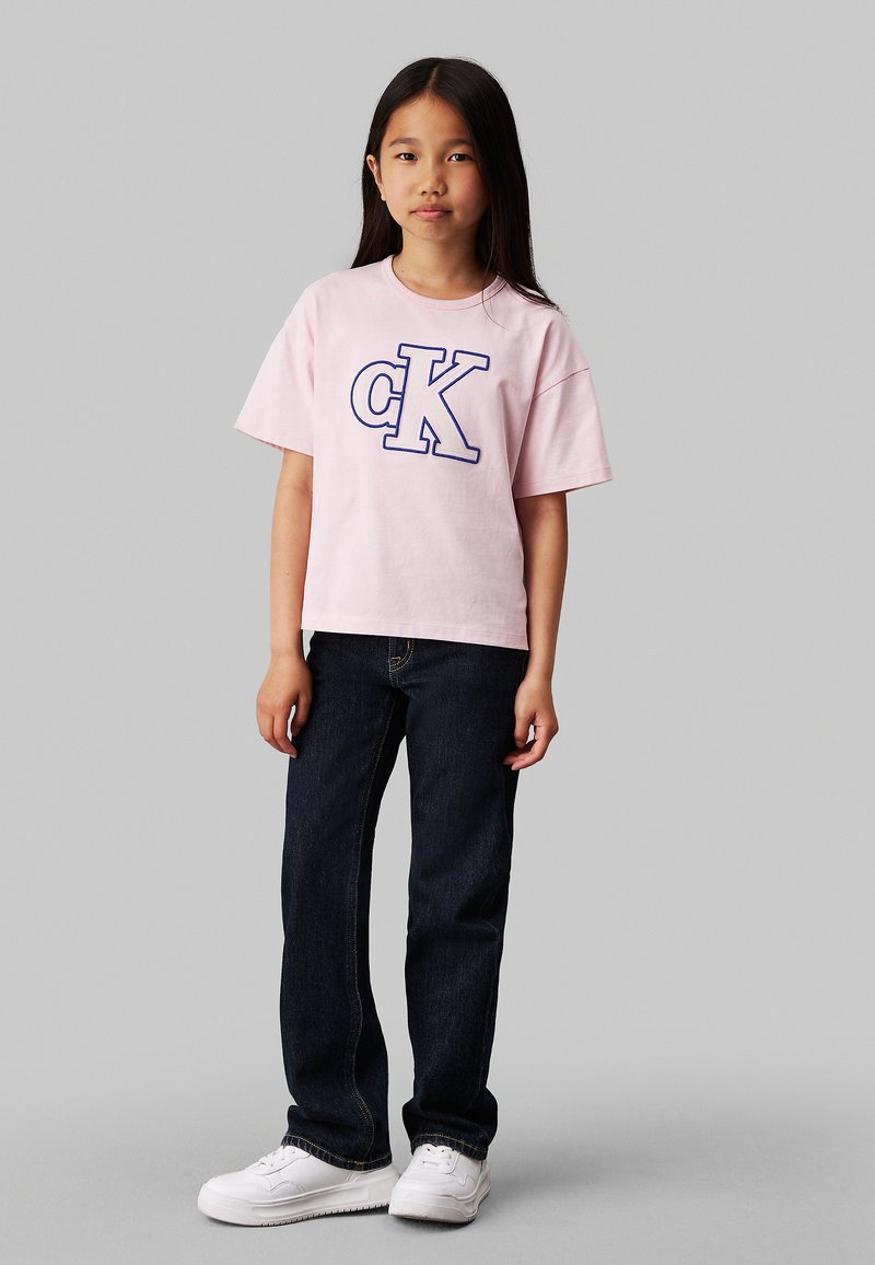 T-shirt in cotone rosa con logo "CK" blu, abbinata a jeans in denim blu scuro e scarpe da ginnastica bianche. Vestibilità comoda e maniche corte.