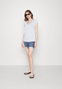 Camiseta azul clara em forma de V com mangas curtas, combinada com uma saia mini de jeans desfiada e chinelos bege, em uma modelo contra um fundo branco.