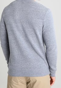Uomo che indossa un maglione grigio chiaro a maniche lunghe e pantaloni beige, mostrato di spalle su uno sfondo chiaro e semplice.