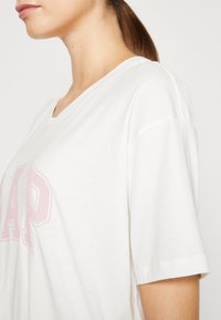 Camiseta blanca de algodón con escote en V y mangas cortas, presenta un logo "GAP" en color rosa claro en el frente. Textura suave, diseño casual.