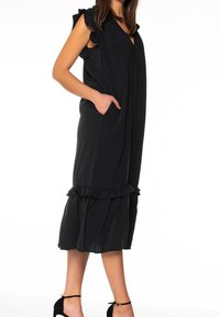 Musta ilman hihoja mekko, jossa on röyhelökoristeita, syvä V-kaula-aukko ja sivutaskut; sisältää kerroksellisen helman röyhelöillä ja rentoa istuvuutta.