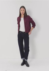 Burgundy bomber-takki, valkoinen t-paita, tummat farkut käärittyinä lahkeista ja mustat saappaat, käytetty studioympäristössä valkoista taustaa vasten.