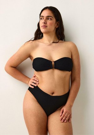 Kvinde med langt mørkt hår iført en tekstureret sort bandeau-bikinitop og bikinitrusser med høj talje, stående med den ene hånd på hoften.