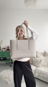 Fehér pulóvert és fekete nadrágot viselő nő egy nagy bézs színű vászontáskát tart a kezében egy nappaliban, ahol kanapé és zöld színes asztal található.