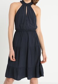 Navy blue halter dress, valmistettu kevyestä kankaasta, jossa on avainaukko-kaula-aukko, muotoiltu vyötärö ja kerroksellinen frilla-helma.