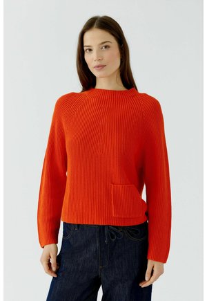 Vrouw met lang bruin haar die een fel oranje gebreide trui met een voorzak en donkerblauwe spijkerbroek draagt tegen een effen lichte achtergrond.