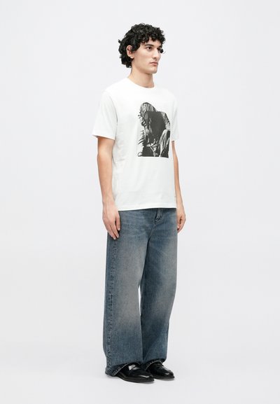 Λευκό βαμβακερό T-shirt με μαύρο γραφικό τύπωμα, σε συνδυασμό με ανοιχτό μπλε τζιν παντελόνι με φαρδιά μπατζάκια και μαύρα δερμάτινα παπούτσια.