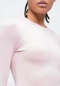 Close-up van een persoon die een lichtroze shirt met lange mouwen draagt, met gevlochten haar zichtbaar tegen een effen witte achtergrond.