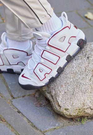 Białe i bordowe sneakersy Nike Air z dużym napisem "AIR", noszone z białymi skarpetkami na kamiennej powierzchni na zewnątrz.