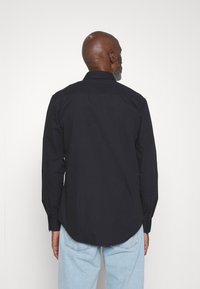 Camisa de botones de un oscuro color azul marino con cuello, mangas largas y un dobladillo curvado, hecha de una suave tela de algodón. Costuras blancas visibles que la acentúan.