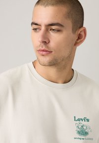 T-shirt de algodão cor creme com logótipo verde da Levi's e design gráfico no lado esquerdo do peito. Mangas curtas e gola redonda.