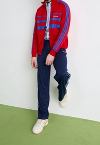 Červená atletická bunda na zip s modrými pruhy, doplněná tmavě modrými sportovními kalhotami s bočními pruhy. Celý outfit dokončují krémové tenisky.