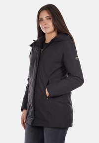Giacca nera isolata con cappuccio, progettata con chiusura frontale a zip, tasche laterali e una texture liscia. Caratterizzata da un badge con logo sulla manica.