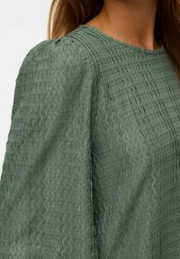 Blouse verte froissée en tissu texturé, avec un col rond et des manches bouffantes. La surface présente des lignes verticales subtiles.