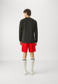 Schwarzes langärmliges Shirt, rote Shorts, weiße kniehohe Socken mit schwarzen Streifen und weiße Fußballschuhe, von hinten betrachtet. Einfaches, sportliches Design.