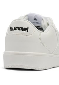 Vita sneakers i slät syntetiskt material med en texturerad sula, med en svart "hummel"-logotyp nära hälen och en minimalistisk design.