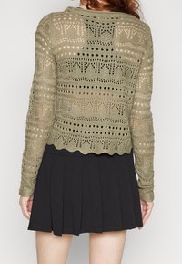 Olivegrüner Strickcardigan mit einem offenen Muster, langen Ärmeln und gewelltem Saum, getragen über einem schwarzen Faltenrock.