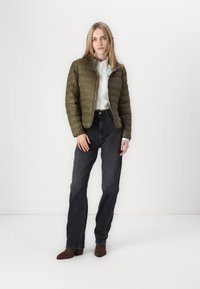 Padded olivově zelená bunda s vysokým límcem, předváděná s bílou košilí na knoflíky a tmavými džíny s vysokým pasem. Hnědé kotníkové boty doplňují vzhled.