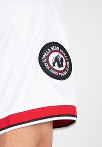 Camiseta atlética blanca que presenta un parche redondo negro con un logotipo de gorila, acentos rojos y un ribete negro y rojo en las mangas y el dobladillo.