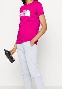 Camiseta fucsia con logo blanco, combinada con pantalones deportivos gris claro. Modelo sosteniendo una botella de agua negra. Conjunto de ropa deportiva simple y casual.