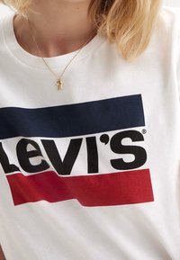 Camiseta de algodón blanca con un audaz logo negro de "Levi's", acentuado por rayas horizontales azules y rojas debajo del logo; textura de tela suave.