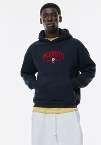 Hanorac bleumarin cu text roșu "Peanuts Golf Team". Are un buzunar frontal și manșete cu nervuri. Purtați peste o cămașă galbenă și pantaloni de trening gri.