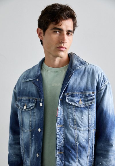 Light blue denim jacket with a classic cut, two chest pockets, and silver buttons, layered over a muted green t-shirt.