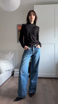 Zwart top met lange mouwen en een wikkelontwerp, gecombineerd met blauw denim spijkerbroek met hoge taille en wijde pijpen, en puntige zwarte hakken.