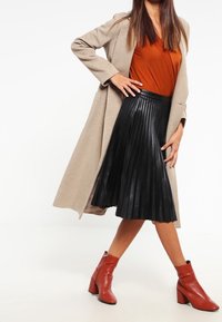 Cappotto beige, top color ruggine, gonna midi plissettata nera e stivaletti rossi con tacco a blocco. Texture morbide e colori contrastanti.