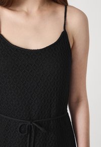 Černé šaty s propracovaným květinovým krajkovým vzorem, přiléhavým korzetem, tenkými ramínky a nastavitelným páskem v pase pro personalizaci.