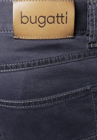 Ciemnoniebieskie spodnie dżinsowe z jasnobrązową skórzaną metką z nazwą marki „bugatti”. Zawierają szczegółowe przeszycia i tradycyjny fason kieszeni.