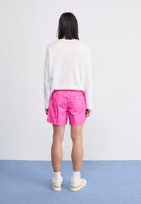 Hvid langærmet skjorte, cropped ved taljen, parret med lyserøde shorts. Sneakers med hvide sokker. Baggrunden har et lysblåt område.