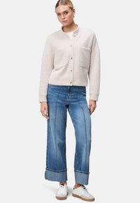 Lys beige, struktureret cardigan med knaplukning og to brystlommer, parret med brede blå denimjeans og hvide sneakers.