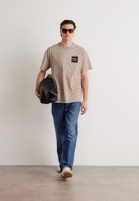 Tricou din bumbac bej cu o mică etichetă logo neagră, purtat cu blugi albaștri și adidași albi. Modelul ține o geantă neagră.