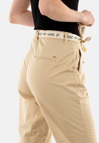 Beige broek met een gladde textuur, voorzien van een brede tailleband met tekst en een zijzak. Het taps toelopende ontwerp benadrukt de vorm.