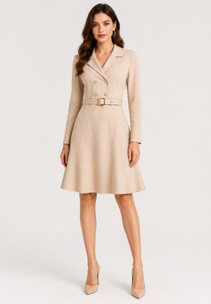 Vrouw draagt een beige, op maat gemaakte jurk met lange mouwen, dubbele knopenrij en een bijpassende riem, gecombineerd met beige hoge hakken.