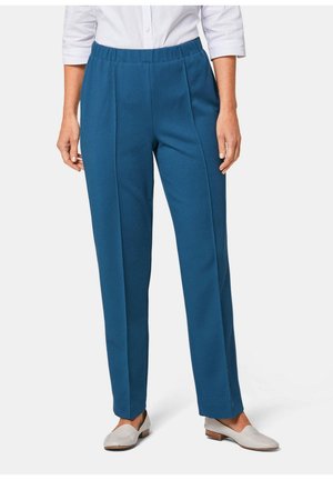 Femme portant un pantalon droit couleur sarcelle avec taille élastique, associé à des mocassins blancs et une chemise blanche boutonnée.