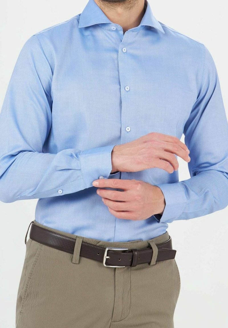Camicia a maniche lunghe azzurro chiaro con colletto a bottoni e bottoni bianchi. Indossata con pantaloni color khaki e una cintura in pelle marrone.