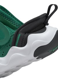 Gros plan du talon d’une chaussure de sport verte et noire avec une semelle blanche texturée et une languette sur fond blanc.