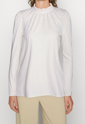 Kobieta ubrana w białą bluzkę z długim rękawem, z marszczonym wysokim kołnierzem i beżowe spodnie na jednolitym tle.