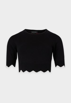 Femme debout, les mains dans les poches, portant un crop top noir à bord festonné, une jupe en jean patchwork et des ballerines noires, sur fond uni.