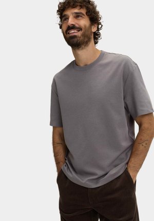 Uomo sorridente con capelli ricci che indossa una camicia grigia a maniche corte e pantaloni di velluto a coste scuri, con le mani nelle tasche, in piedi davanti a uno sfondo semplice.