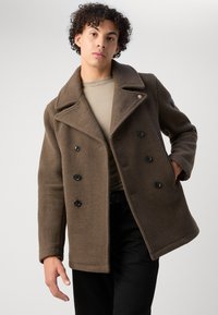 Brun ull peacoat med doble knapper, stor flat krage, knappdetaljer og sidelommer, kombinert med en beige topp og svarte bukser.