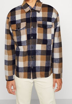 Hombre con camisa de cuadros marrón, negro, blanco y gris, con dos bolsillos en el pecho y pantalones de color crema, posando sobre un fondo blanco.