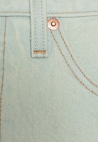 Tela de denim azul claro con costuras visibles y un detalle de remache de cobre, con un acabado texturizado y un patrón de tejido consistente.