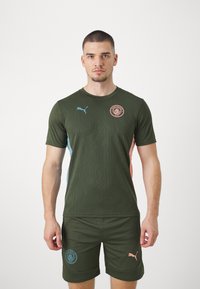 Zaļš sporta krekls ar teksturētu rakstu, ziliem un rozā sānu paneļiem, Manchester City logos, apvienots ar zaļiem šortiem un Puma akcentiem.