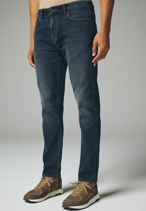 Donkerblauwe denim jeans met een slim fit, voorzien van vijf zakken, een ritssluiting en contrasterend stiksel. G gedragen met bruine sneakers.