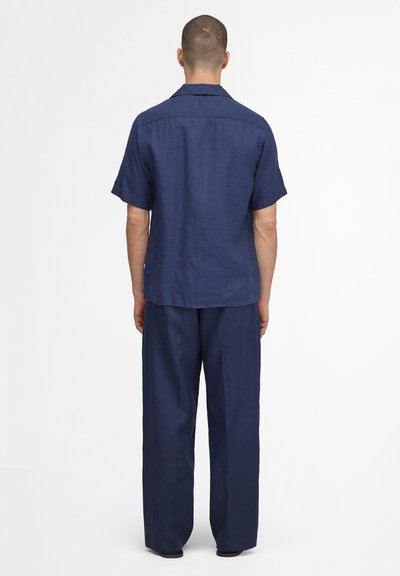 Homme debout de dos, portant une chemise à manches courtes bleu foncé et un pantalon ample assorti sur un fond blanc uni.