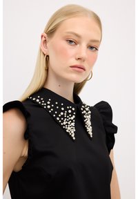Sort top med korte flæseærmer og en krave prydet med hvide perler og klare krystaller, med en blød stoftekstur.